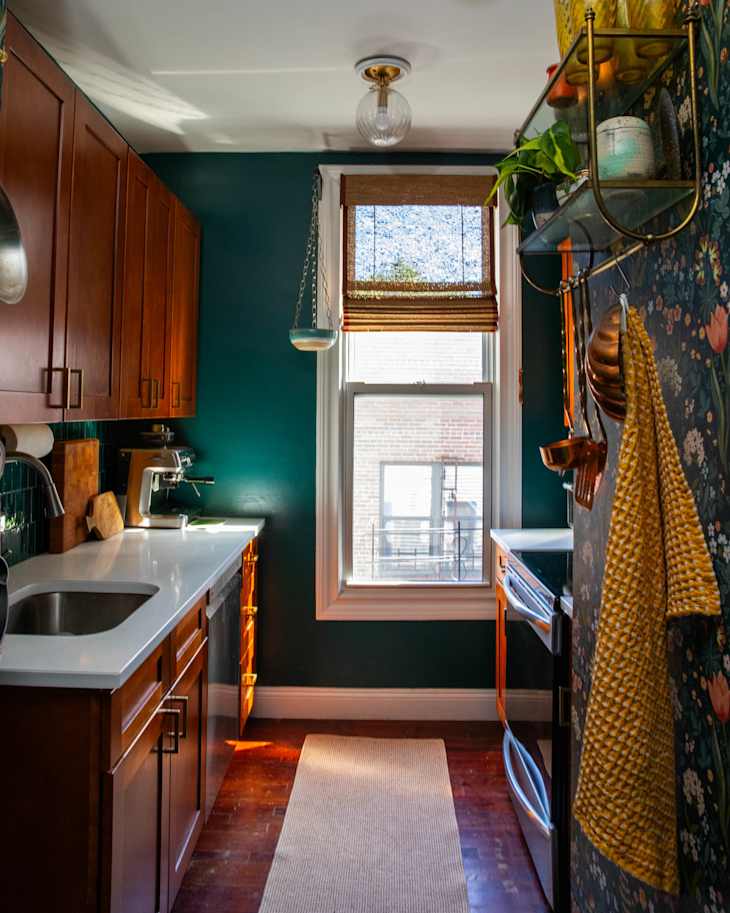 Cozy kitchen with dark green walls, wooden cabinets, white countertops, and a window with bamboo shades.