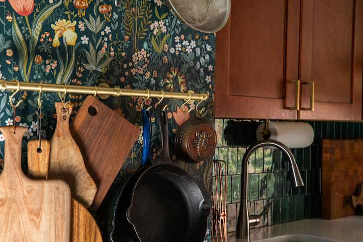 Wooden cutting boards and cast iron skillet hanging on a gold rack against floral wallpaper in a kitchen.