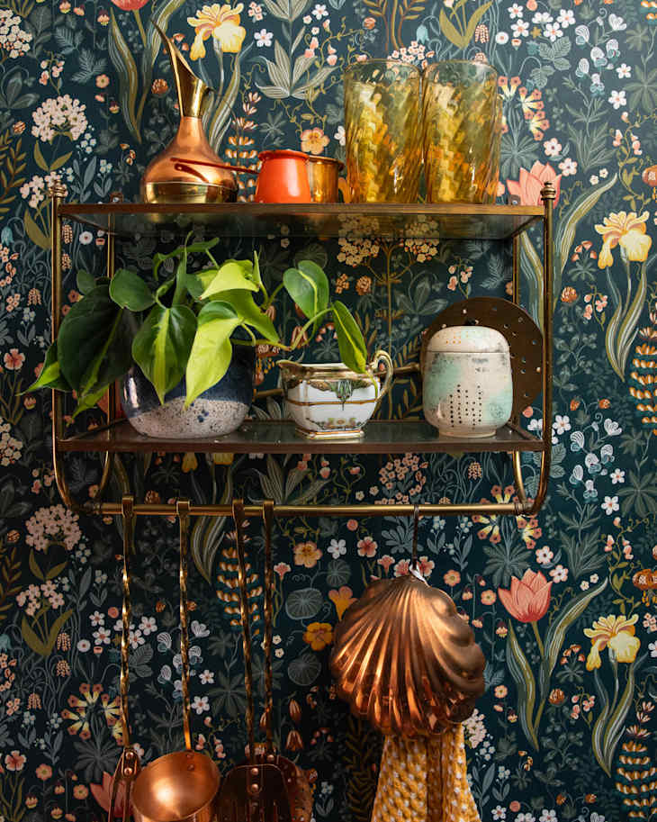 Brass shelf with colorful vases, a plant, and kitchen utensils against a floral wallpaper backdrop.