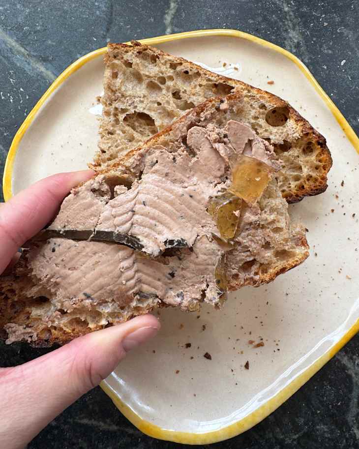 A hand holding a slice of bread topped with pâté and garnished with jelly, resting on a cream-colored plate.