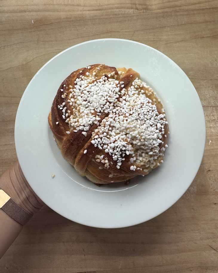 A hand holding a white plate with a sweet pastry topped with sugar crystals, resting on a wooden surface.