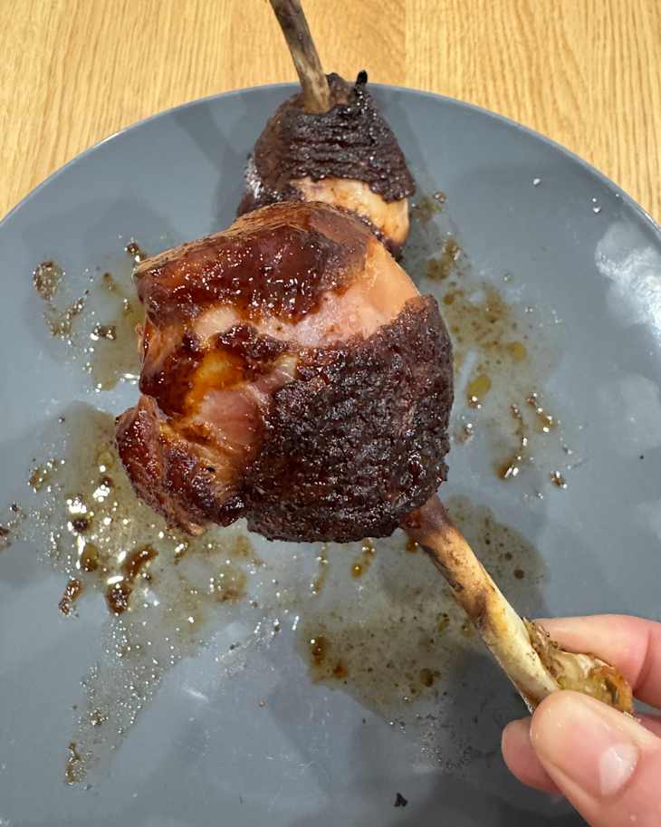 Two smoked meat drumsticks on a gray plate, with a hand holding one and sauce drips underneath.
