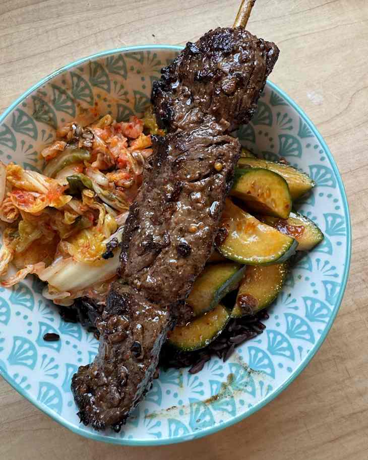 Grilled beef skewer on a patterned plate with kimchi, sautéed zucchini, and black rice.