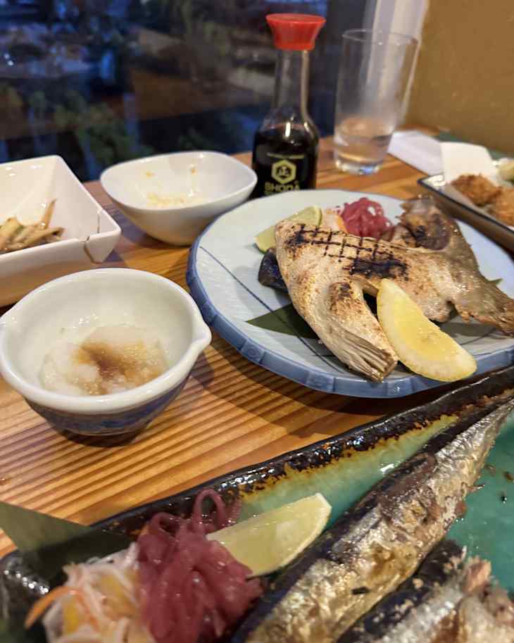 Grilled fish served with lemon wedges, pickled vegetables, dipping sauce, and a side of grated daikon on a wooden table.