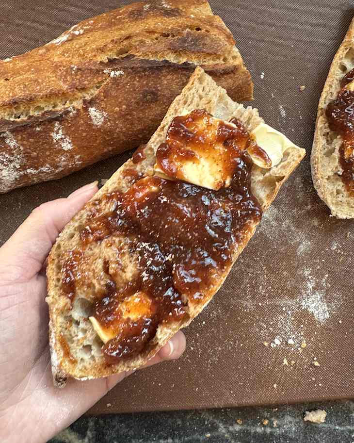 A hand holding a slice of bread topped with butter and fruit jam, with a loaf of bread in the background.