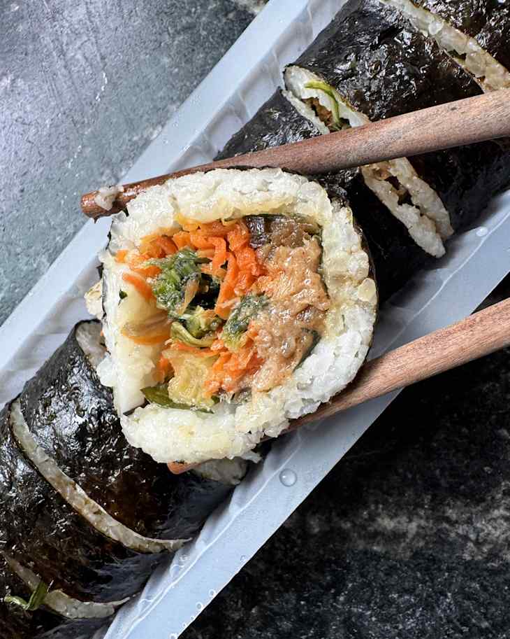 Sliced sushi roll with vegetables, held by chopsticks, on a gray surface.