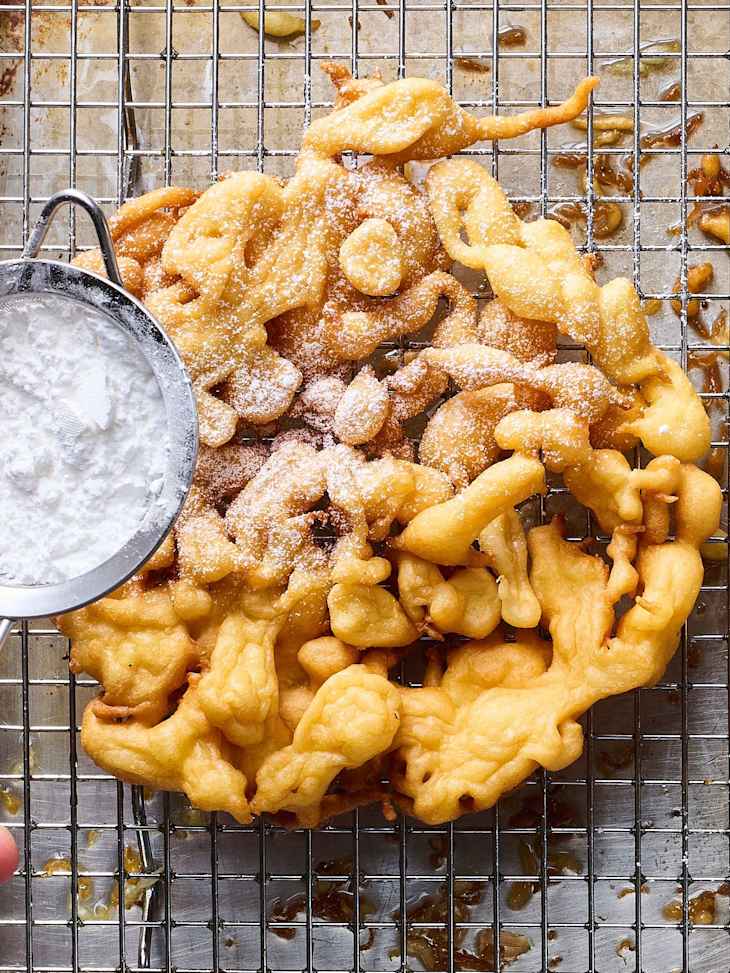 Fried funnel cakes dusted with powdered sugar on a wire rack, with a hand holding a sieve.