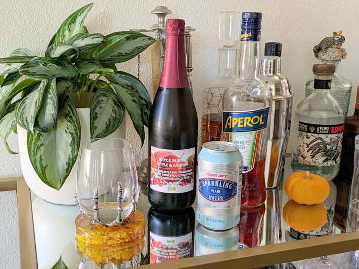 A decorative bar cart featuring a green plant, a bottle of sparkling apple and lingonberry drink, Aperol, a shaker, and a can of sparkling water, with a small pumpkin.
