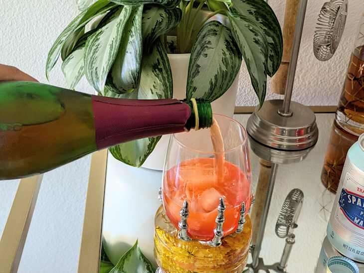 A hand pouring sparkling wine into a glass with a pink drink, surrounded by a plant and decorative items.