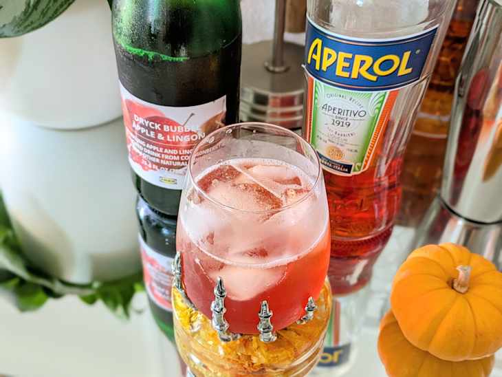 A glass of pink drink with ice, Aperol bottle, and a small orange pumpkin on a reflective surface.