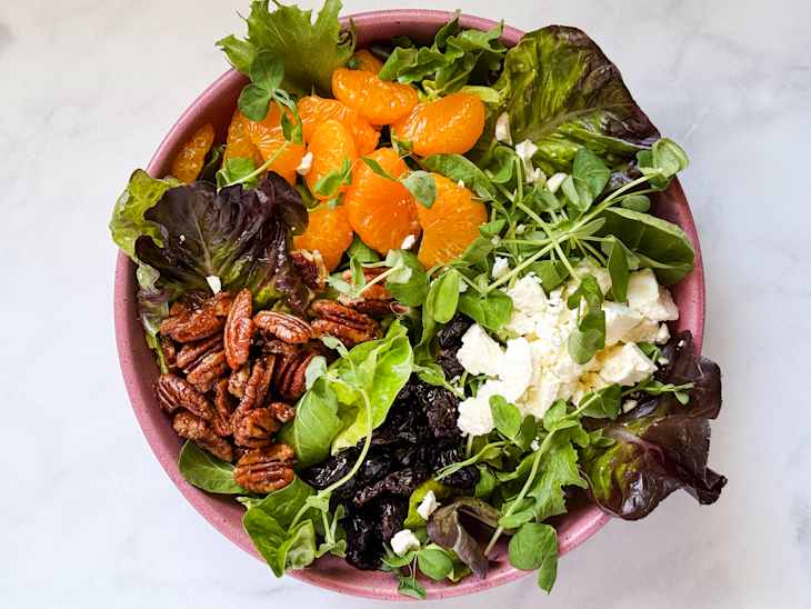 A colorful salad in a pink bowl featuring mixed greens, mandarin oranges, pecans, goat cheese, and pea shoots.