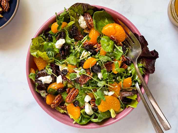 A colorful salad in a pink bowl featuring mixed greens, mandarin oranges, pecans, dried cherries, and crumbled feta cheese.
