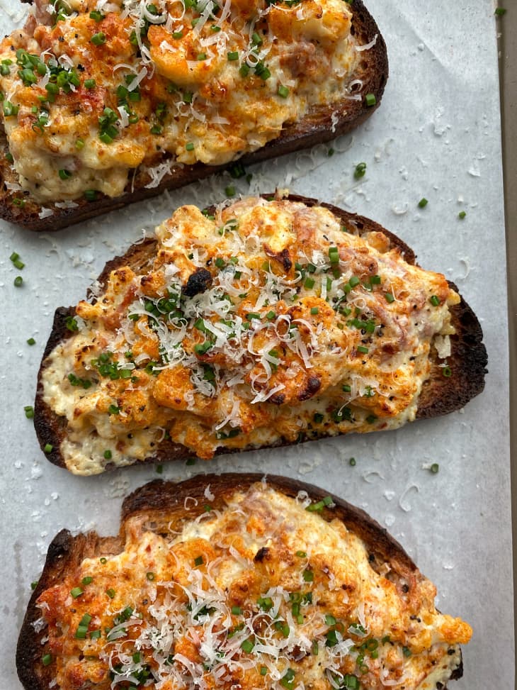 Three slices of toasted bread topped with cheesy cauliflower, chives, and grated cheese on parchment paper.