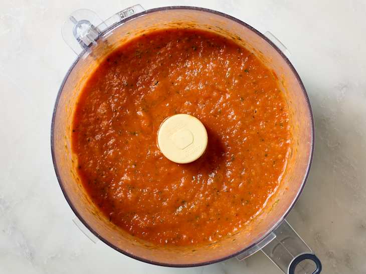 Blended tomato sauce with herbs in a food processor bowl on a light surface.