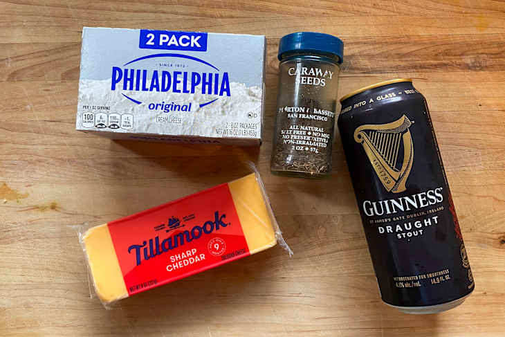 Cream cheese, sharp cheddar cheese, caraway seeds, and a can of Guinness stout on a wooden surface.