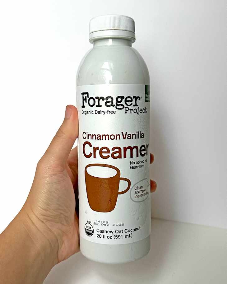 A hand holding a bottle of Forager Project Cinnamon Vanilla Creamer, featuring a white label and a coffee cup illustration.