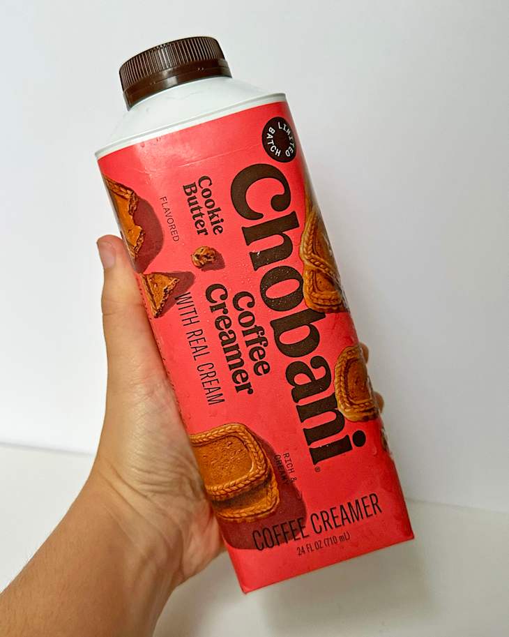 A hand holding a Chobani Cookie Butter coffee creamer container with a red label and brown cap.