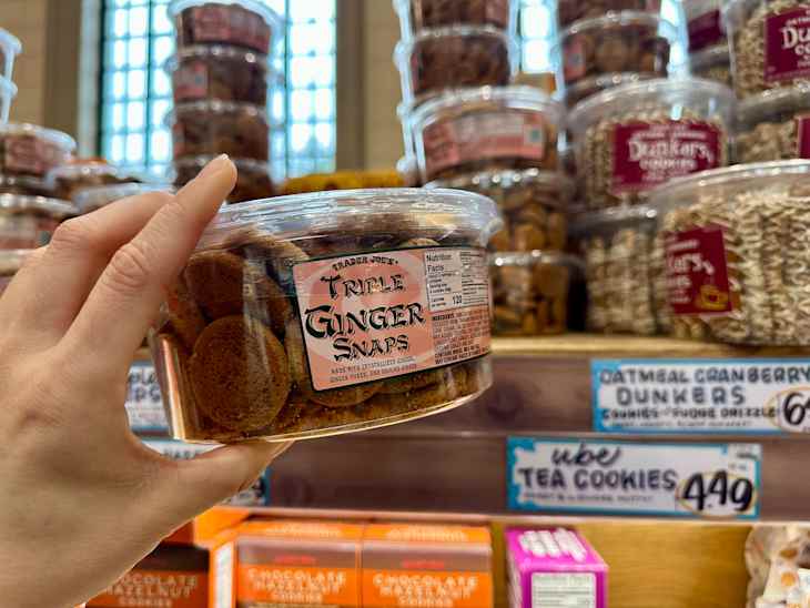 A hand holding a container of Trader Joe's Triple Ginger Snaps in a grocery store aisle with various cookie jars in the background.