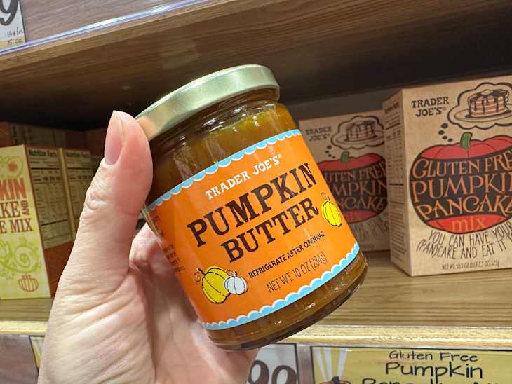A hand holding a jar of Trader Joe's Pumpkin Butter in a grocery store aisle, with pumpkin pancake mix boxes in the background.