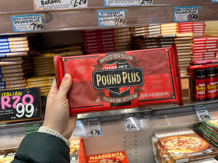 A hand holding a Trader Joe's Pound Plus dark chocolate bar in a grocery store aisle with various chocolate bars and products.