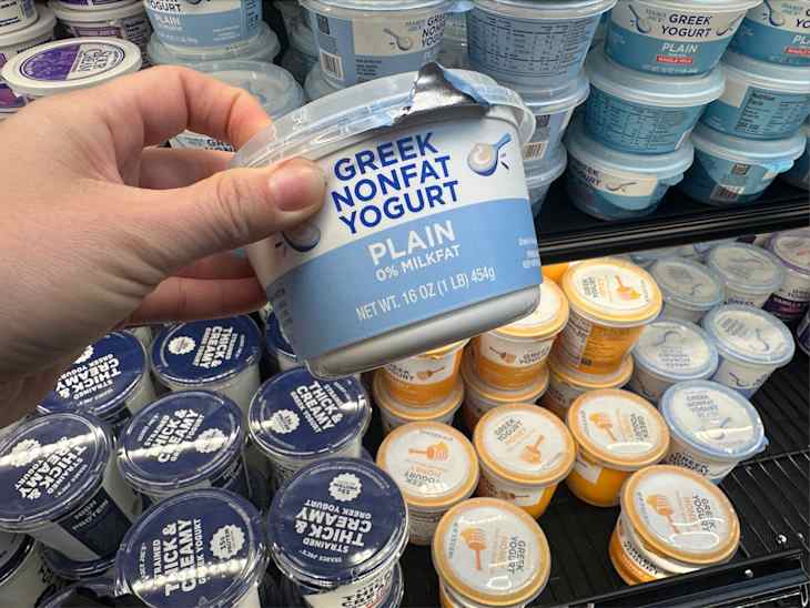 A hand holding a container of Greek nonfat yogurt labeled "Plain" in a dairy section filled with various yogurt containers.