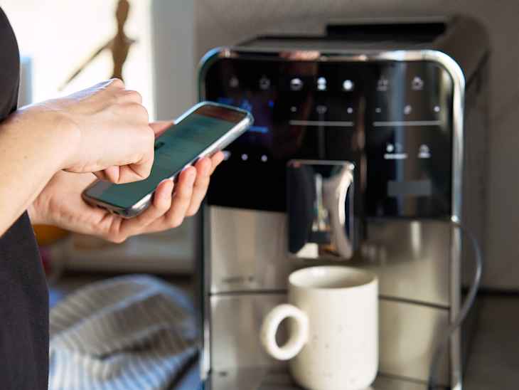 Person uses smartphone app to operate coffee machine. Woman preparing fresh cup of coffee with modern espresso machine