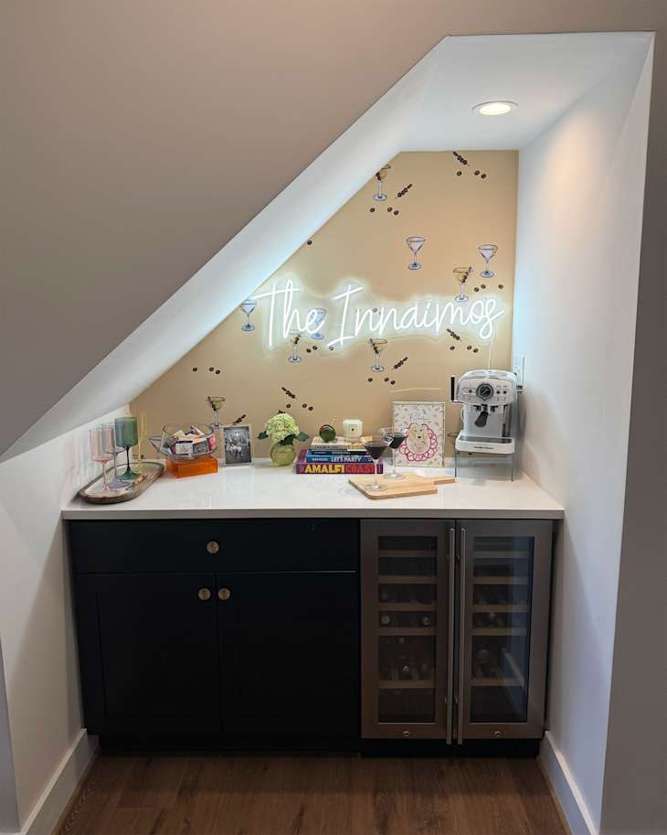 Neon sign mounted on martini motif wallpaper in home bar with books, glasses and barware on top of white countertop.