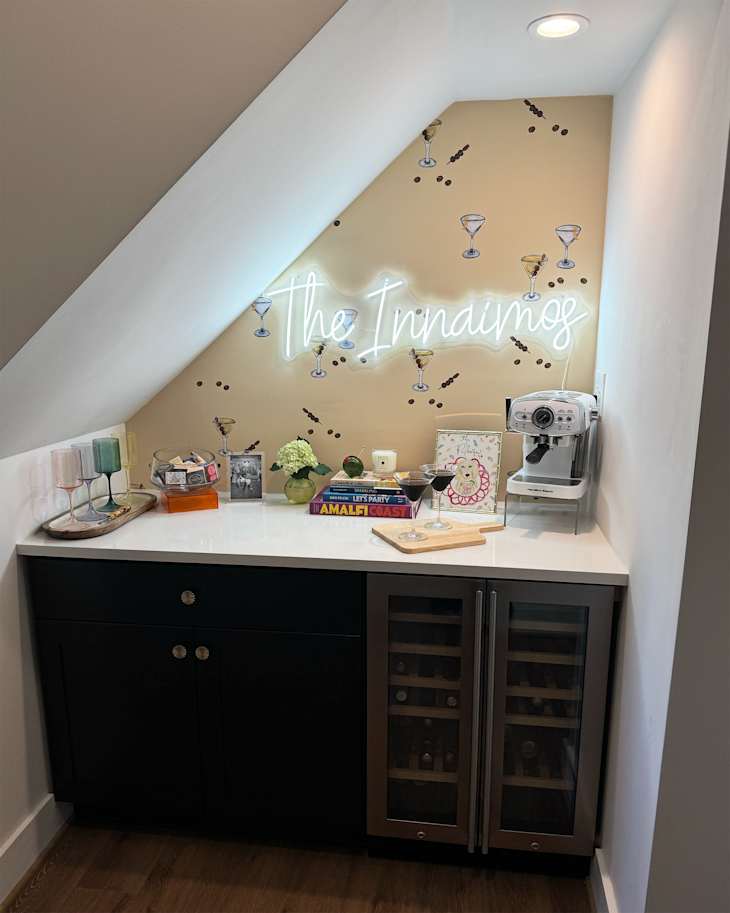 Neon sign mounted on martini motif wallpaper in home bar with books, glasses, espresso maker and barware on top of white countertop.