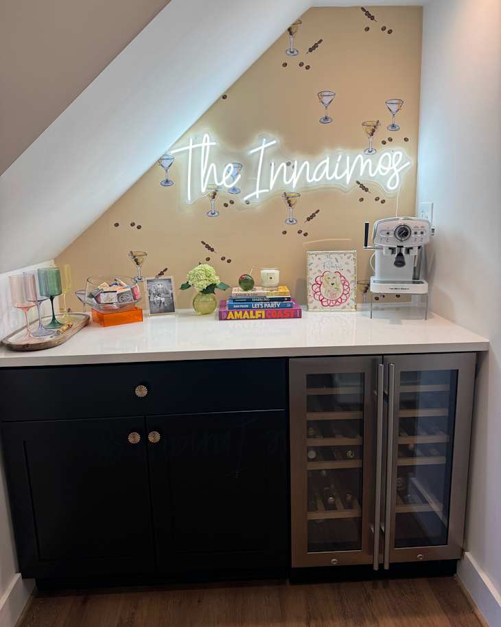 Neon sign mounted on martini motif wallpaper in home bar with books, glasses, espresso maker and barware on top of white countertop.