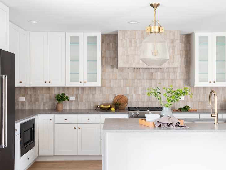A kitchen detail with a grey stone countertop island, gold faucet, stainless steel appliances, brown tile backsplash, and white cabinets. No brands or labels.