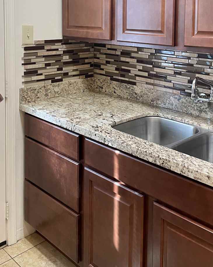 Neutral toned granite countertops in kitchen with brown tile backsplash and dark wooden cabinets before renovation.