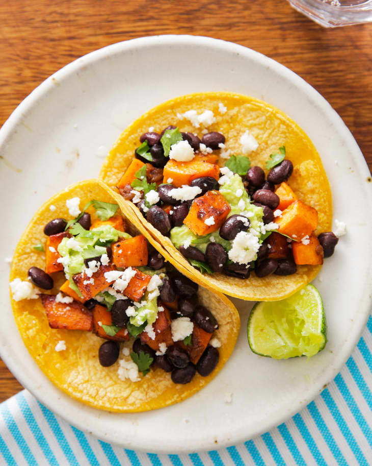 Two tacos with black beans, sweet potatoes, avocado, and cheese on a plate with lime wedge.