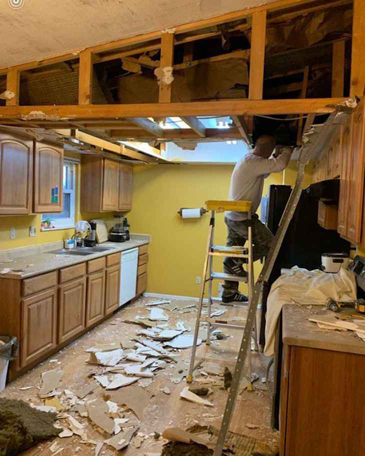 A person on a ladder working on a kitchen ceiling, with debris scattered on the floor and yellow walls.