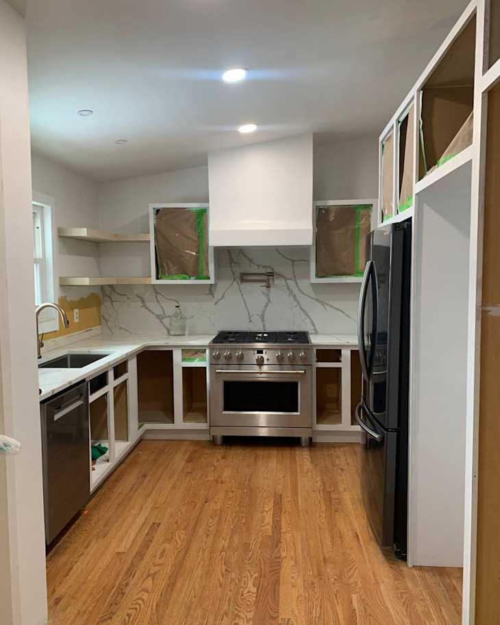 Modern kitchen under renovation featuring a stainless steel stove, dark refrigerator, and white cabinetry with marble backsplash.