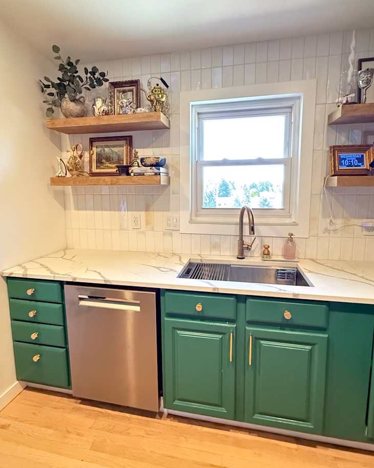 Modern kitchen with green cabinets, stainless steel dishwasher, marble countertop, and open shelves displaying decor and plants.