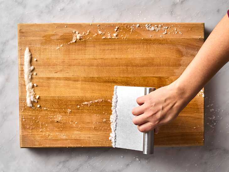 A hand using a metal bench scraper to clean a wooden cutting board with scattered salt.