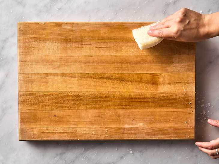 A wooden cutting board being cleaned with a sponge, with a hand visible in the process.