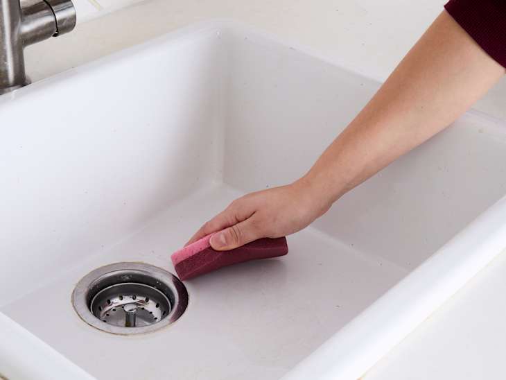 A hand scrubbing a stainless steel sink drain with a pink sponge, showing a clean white sink.