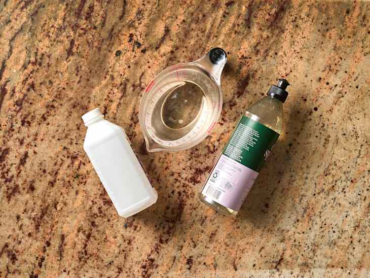 A white plastic bottle, a clear measuring cup with water, and a green liquid bottle on a granite countertop.