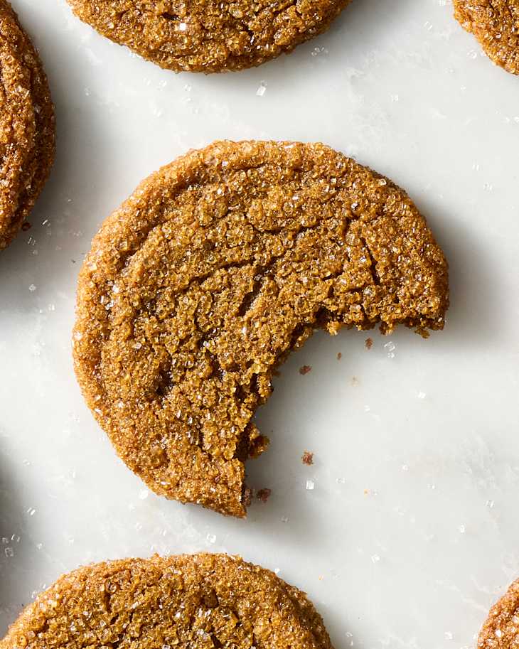 Molasses cookies with visible sugar crystals on a light surface with one cookie bitten into.
