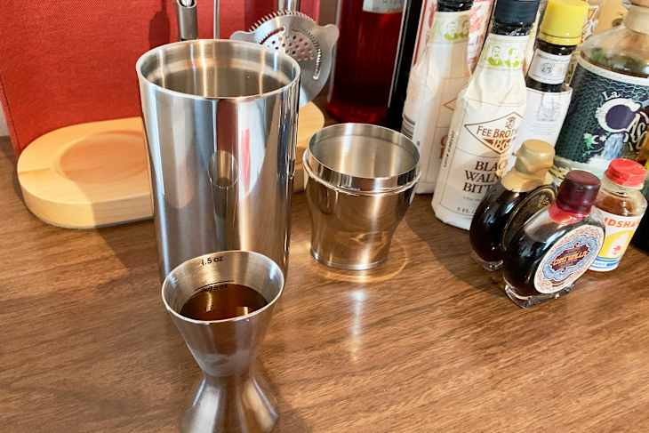 Stainless steel cocktail shaker with ice, strainer, and various bottles.