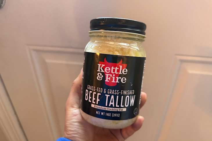 A person holding a jar of beef tallow labeled "Kettle & Fire," with a black lid and a clear glass container.