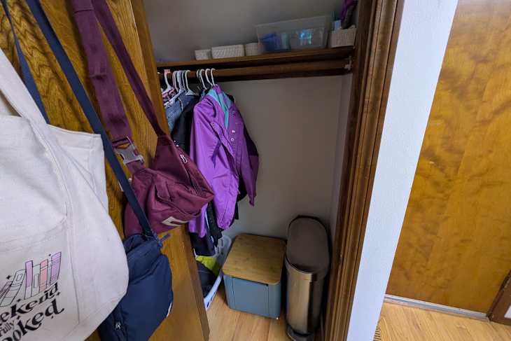 Closet with hanging jackets, bags, a laundry basket, a blue storage box, and a silver trash can.