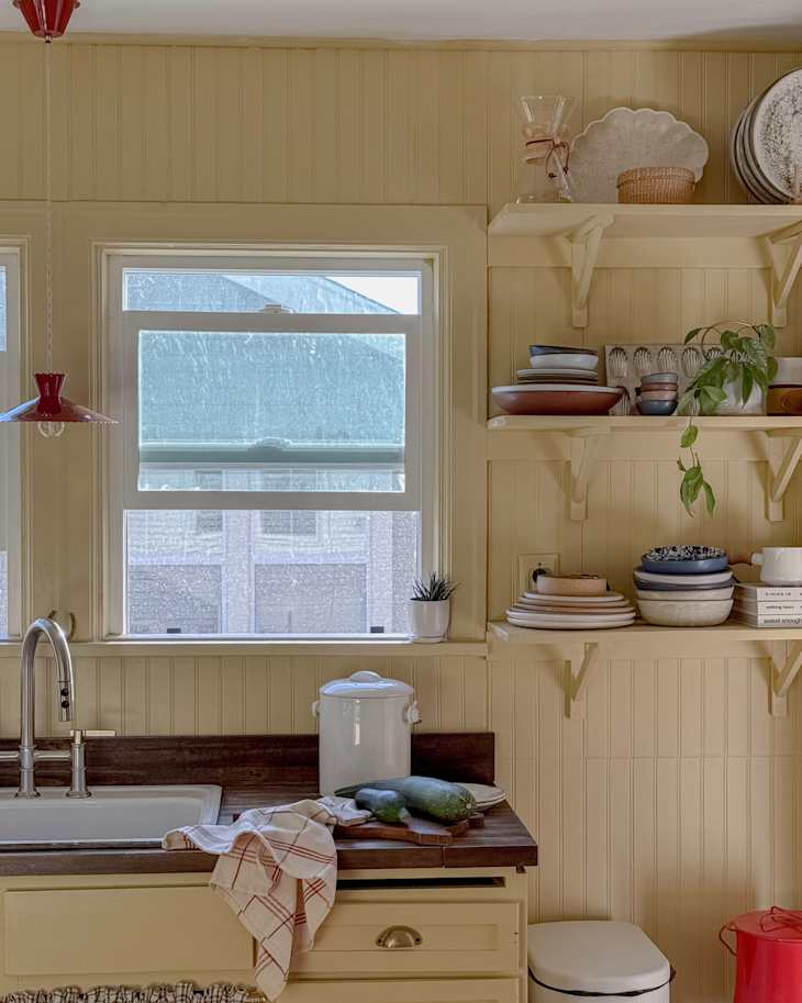Bright yellow kitchen with a sink, wooden countertop, fresh zucchini, and open shelves displaying various dishes and plants.