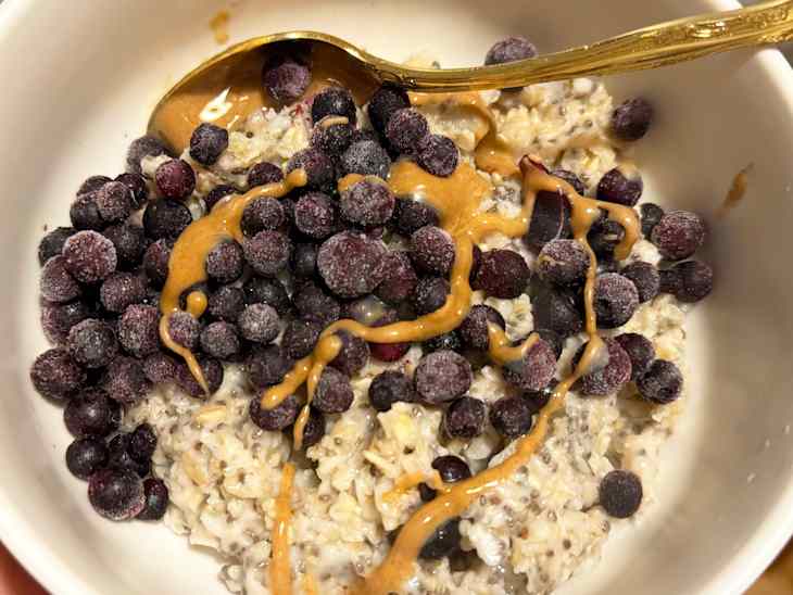 Bowl of oatmeal topped with frozen blueberries and drizzled with nut butter, with a gold spoon resting inside.