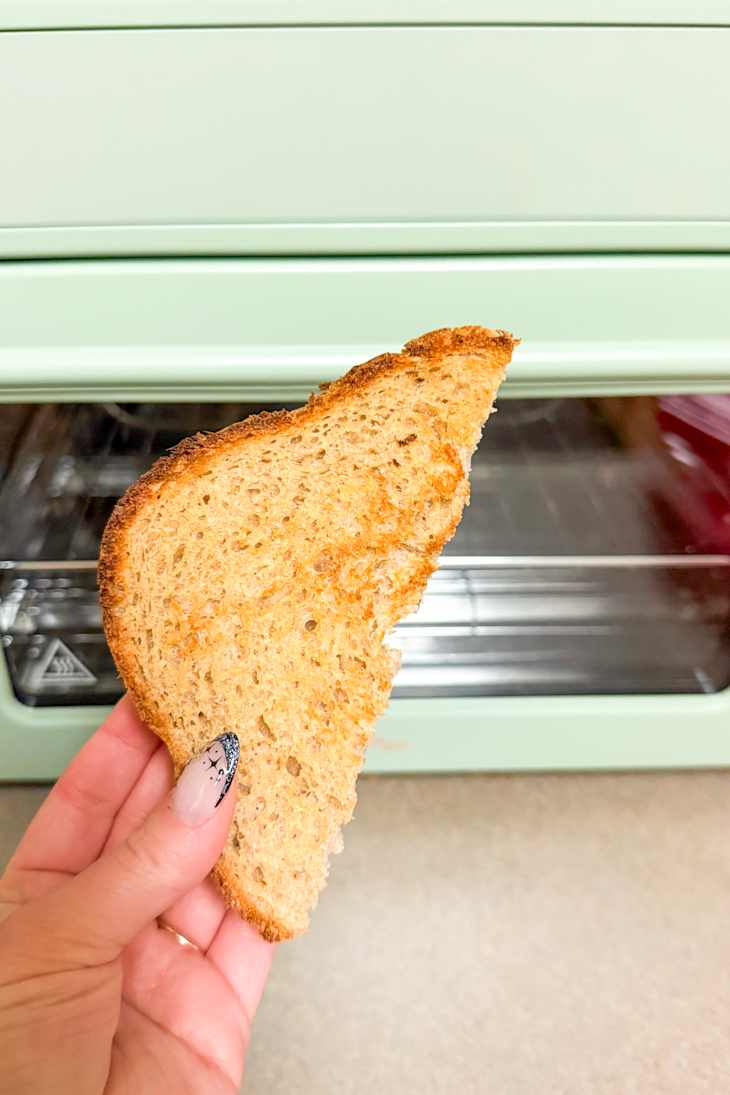 A hand holding a slice of toasted whole grain bread in front of a mint green air fryer.