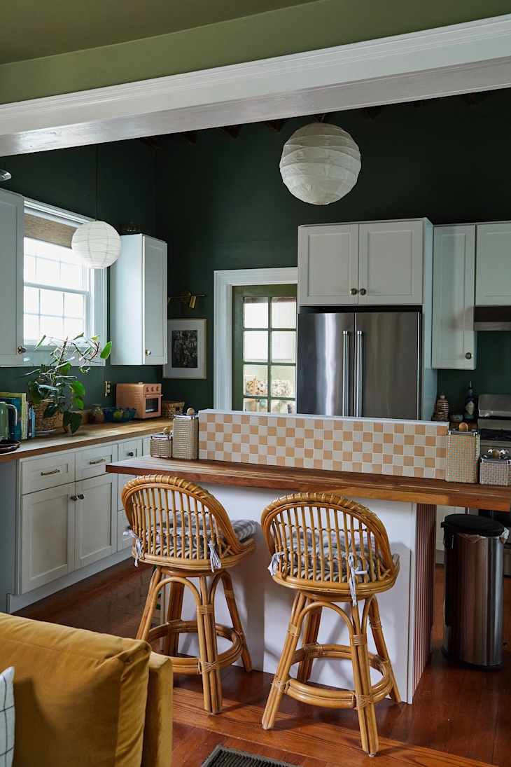 Cozy kitchen with dark green walls, white cabinets, rattan bar stools, and a patterned backsplash.