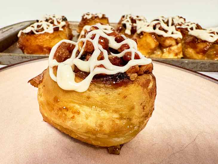 Cinnamon roll topped with icing and nuts, placed on a light plate with more rolls in the background.