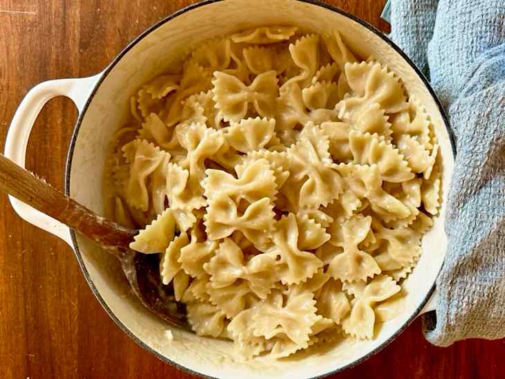 Bowed pasta in a white pot with a wooden spoon, next to a blue kitchen towel on a wooden surface.