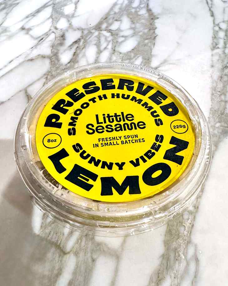 Container of preserved lemon hummus with a bright yellow label on a marble surface.